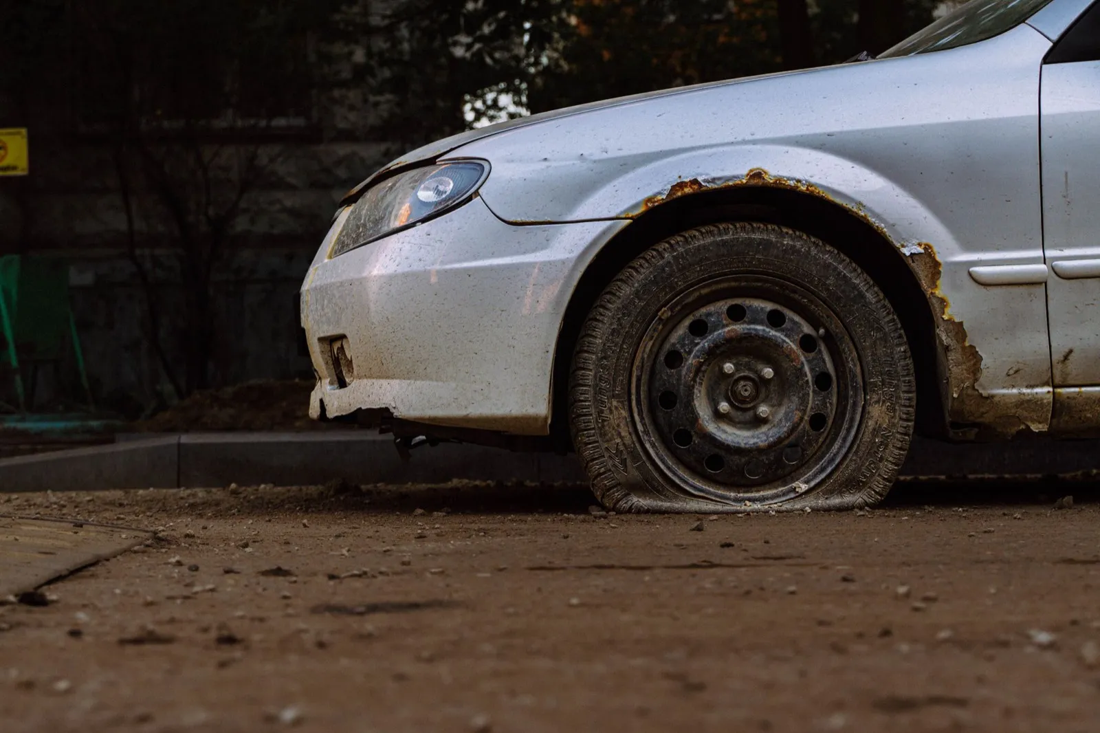A rusted silver sedan with a flat front tire, left parked at a curb